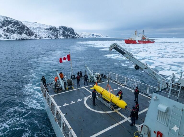 Naval personnel preparing a yellow autonomous underwater vehicle (AUV) for deployment on a ship deck in an icy Canadian Arctic environment, featuring a Canadian flag and a red research vessel in the background | The Deltic Group